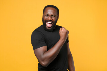 Wall Mural - Joyful african american man guy football fan in casual black t-shirt isolated on yellow background studio portrait. People sincere emotions lifestyle concept. Mock up copy space. Doing winner gesture.