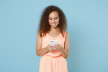 Smiling young african american girl in pastel summer clothes isolated on blue wall background studio portrait. People lifestyle concept. Mock up copy space. Using mobile cell phone typing sms message.