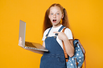 Young redhead school girl 12-13 years old in white t-shirt blue denim uniform backpack hold laptop pc computer isolated on yellow background children studio portrait Kids education lifestyle concept