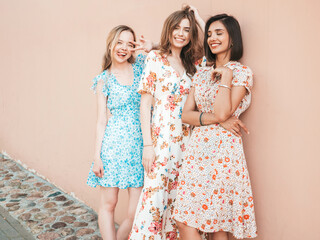 Wall Mural - Three young beautiful smiling hipster girls in trendy summer sundress.Sexy carefree women posing on the street near wall. Positive models having fun and hugging
