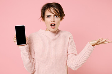 Wall Mural - Perplexed young brunette woman in knitted casual sweater isolated on pastel pink background. People lifestyle concept. Mock up copy space. Hold mobile phone with blank empty screen, spreading hands.