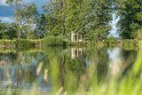 Fototapeta Na ścianę - A view with blurred grass in the foreground across a beautiful lake with reflections of trees towards an old abandoned structure in the middle of a rural countryside scene