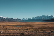 Peaceful Landscape Of Dry Grass Field And Far Away Lake At The Foot Of The Mountain With Snow On The Mountains Top In Patagonia, Chile.