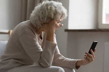 Sad elderly woman sit on sofa hold smartphone feels disappointed by received sms bad news, awful message, difficulties with modern device usage, unpleasant notification, stressed older person concept