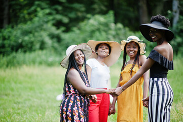 Group of four gorgeous african american womans wear summer hat holding hands and praying at green grass in park.