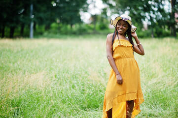 Portrait of gorgeous african american woman 20s in wear in yellow dress and summer hat posing at green grass in park.