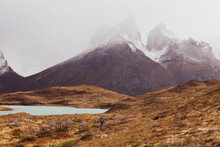 Person Standing Far Away In Peaceful Landscape Of Dry Grass Fields And Small Lake. Background Of Mountains With Snow On Their Summit In Patagonia, Chile.