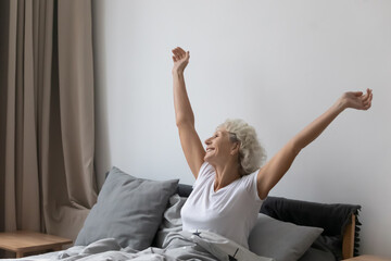 Wall Mural - Optimistic refreshed middle aged senior woman sitting in bed wake up feel rested after enough healthy sleep in comfortable bed, stretching arms smiling looking out the window enjoy and welcome new day