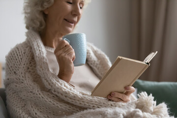 Mature woman wrapped in warm knitted plaid relax on couch drink tea, read favourite book, classic literature, enjoy carefree time on weekend, close up. Pastime and hobby, tranquil retired life concept