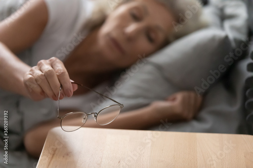 Elderly woman go to bed took off glasses put them on bedside table after evening reading close up view at eyewear and hand. Senior people laser vision eyesight correction, specs offer store ad concept