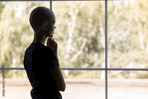 Back view of young sick hairless woman suffer struggle with oncology look in window distance think dream of recovery, ill bald female patient lost in thoughts, feel hopeful, healthcare concept