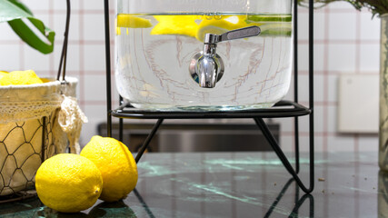 Fresh cold mineral lemon water in a glass tank and two lemons on the green marble table, summer refreshment, detox healthy water, diet plan concept, citrus juice drink 