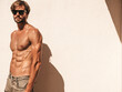 © halayalex - Portrait of sporty handsome strong man. Healthy athletic fitness model posing near white wall in jeans. Confident sexy fashion male with naked nude torso. Lambersexual outdoors in sunglasses