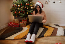 Stylish Young Woman Sitting With Laptop Under Christmas Tree With Lights And Ornaments In Modern Room. Happy Girl In Santa Hat Working On Laptop, Shopping On Christmas Sales.