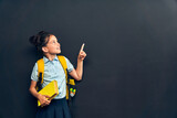 Fototapeta Łazienka - smart girl in uniform with backpack and book in her hand, pointing up