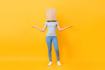 Anonymous woman in casual attire cover head with paper bag doing open hand gesture in yellow studio background