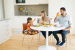 © Mangostar - Family couple and girl having breakfast together in kitchen, sitting at dining table, drinking orange juice and talking. Full length, copy space. Family morning concept