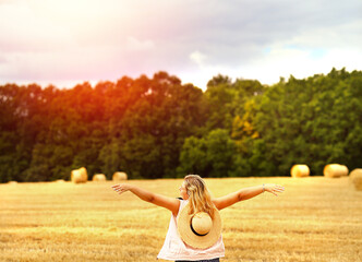 Wall Mural - A young Caucasian blonde woman raised her hands to the sky like wings on a mown wheat field where there is a huge sheaf of hay enjoying nature at sunset. People and travel. Nature. sun rays Agricultur