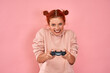 © antianti - Red haired teenage girl has gamble mood plays video games with joystick on pink wall background. Female in pink hoodie has fun at home, holds playstation game controller with interest, feels crazy