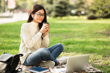 Wall Mural - Girl drinking hot coffee, using laptop, sitting in the park