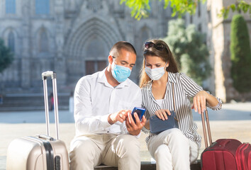 Tourist couple in medical masks to prevent respiratory infection consulting navigation app in cellphone during walk around city..