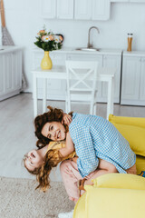 Wall Mural - smiling nanny holding happy kid upside down while having fun on sofa in kitchen