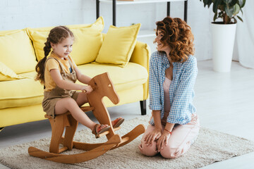 Wall Mural - happy babysitter sitting on floor near adorable child riding rocking horse