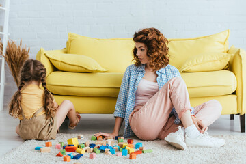 Wall Mural - back view of offended child sitting on floor near young nanny and multicolored blocks