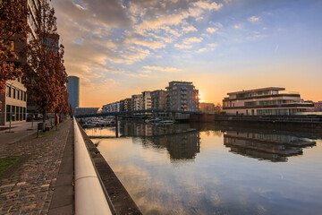 Wall Mural - Canal from the river Main in Frankfurt. Residential area in the morning at sunrise. Water front with reflection. Blue sky and clouds. Shore in the port and buildings with trees and brown leaves