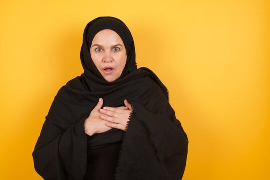 Middle aged muslim woman wearing black hijab over yellow backgro looks with frightened expression, keeps hands on chest, being puzzled to notice something strange,  People, hush reaction and emotions.