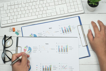 Flat lay, Top view office table desk with businessman hand holding white pen and mouse, document graph analysis and keyboard computer, eyeglass and paper clip