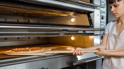 Wall Mural - Young caucasian woman baker is holding a wood peel with fresh pizza and put it in an oven at a baking manufacture factory.