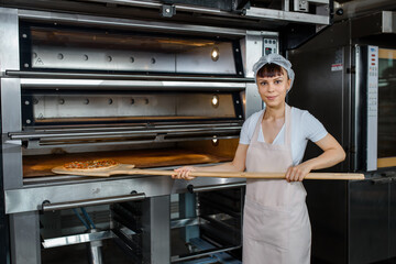 Wall Mural - Young caucasian woman baker is holding a wood peel with fresh pizza near an oven at a baking manufacture factory.