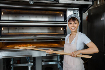 Wall Mural - Young caucasian woman baker is holding a wood peel with fresh pizza near an oven at a baking manufacture factory.