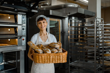 Wall Mural - Young caucasian woman baker is holding a big basket full of fresh bread in the baking manufacture factory.