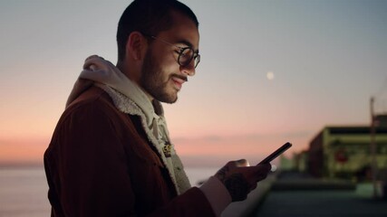 Side portrait of handsome confident young guy in glasses staying online outdoors sunrise, cheerful millennial man influencer using smartphone device smiling connected in social media network