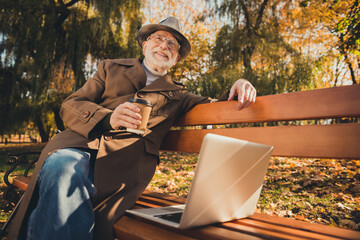 Canvas Print - Photo of white grey hair positive old man enjoy autumn nature outside park rest relax sit bench with takeaway coffee mug use laptop look sunshine wear coat jacket hat cap headwear