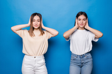 Portrait of two funny mixed race women covering ears isolated over blue background