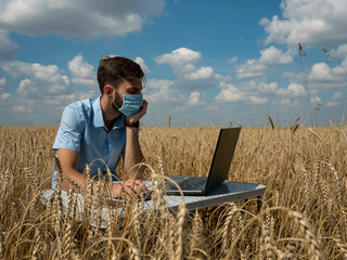 business men at remote work. A young man wearing a Covid-19 mask works for a laptop in a wheat field.