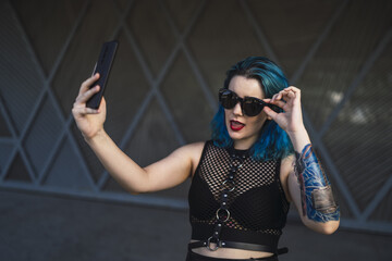 Wall Mural - Cool young lady with blue hair, tattoos, and sunglasses, wearing a crop top taking a selfie