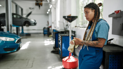 Wall Mural - Doctor for your vehicle. Young african american woman, professional female mechanic looking away, wiping, cleaning her hands with cloth after repairing a car in auto repair shop