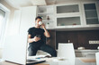 © Strelciuc - Modern caucasian man drinking a coffee while chatting on mobile in the kitchen and working at the laptop