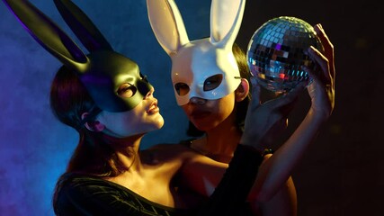 Wall Mural - Portrait of two young flirting cute Asian women in black and white rabbit mask playfully posing for camera with small disco ball on the neon lights - video in slow motion