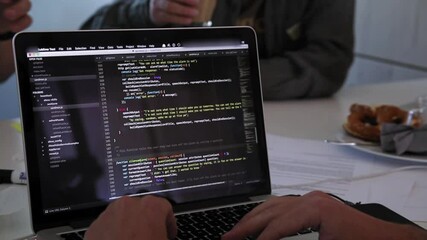 Handheld detail shot of a male person writing code on a laptop. People in the back drinking coffee.