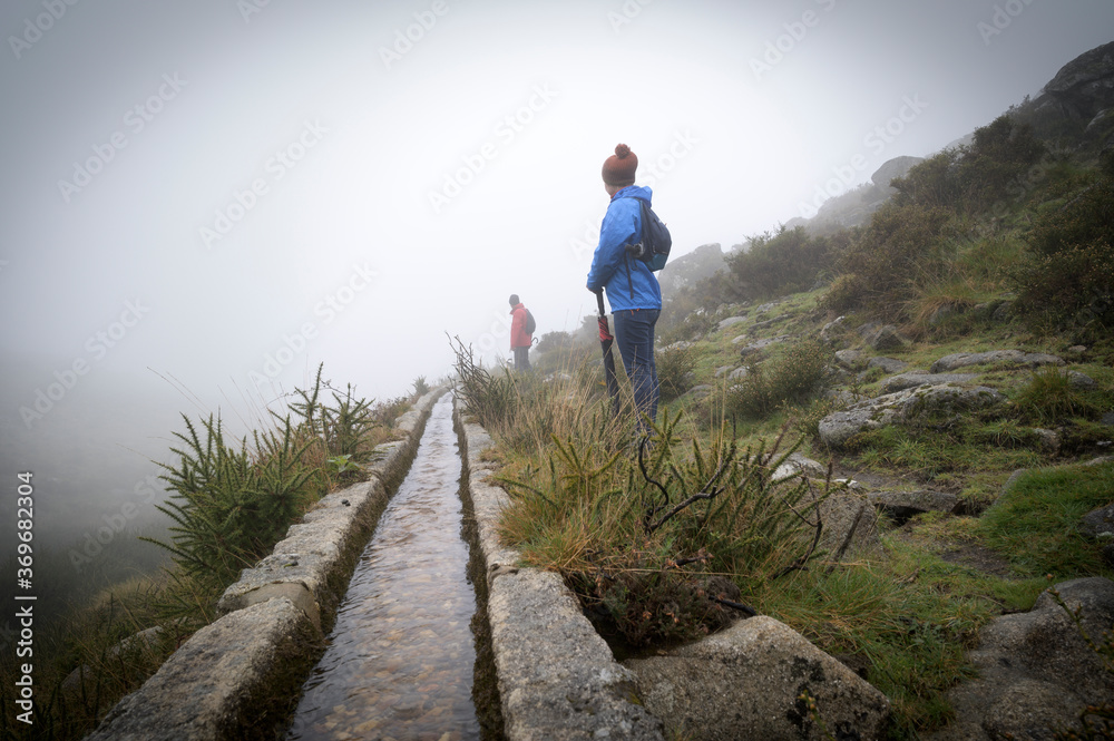 Two people hiking in the mountains in a foggy and rainy day. Girl with blue jacket and umbrella and man with red jacket. - obrazy, fototapety, plakaty 