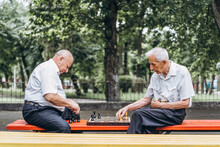 Two Senior Adult Men Playing Chess On The Bench Outdoors In The Park.