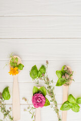 Wall Mural - top view of herbs and green leaves in spoons near flowers on white wooden background, naturopathy concept