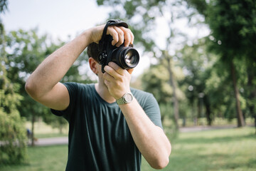 Canvas Print - Front view of man taking photos in nature. Portrait of photographer looking through camera viewfinder and taking pictures while standing in park.