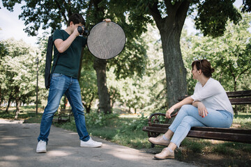 Wall Mural - Male photographer taking photos of girl in nature. Photographer man holding camera and reflector while taking photos of female model in park.