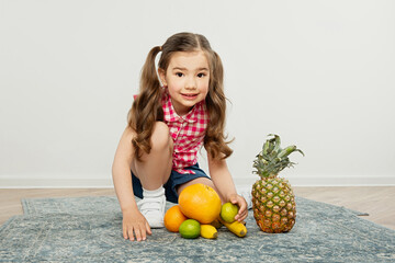 little girl sits on  carpet, juicy fruits lie nearby in heap